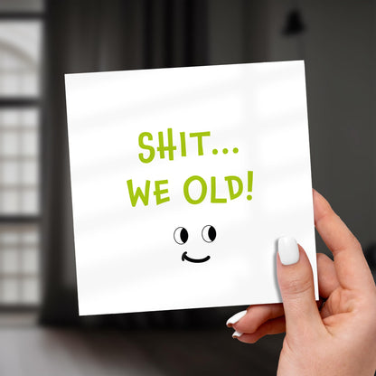 Hand holding a card with 'SHIT... WE OLD!' text and a smiley face on a blurred background
