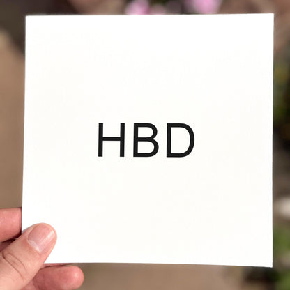 Hand holding a white card with 'HBD' on it against a blurred background