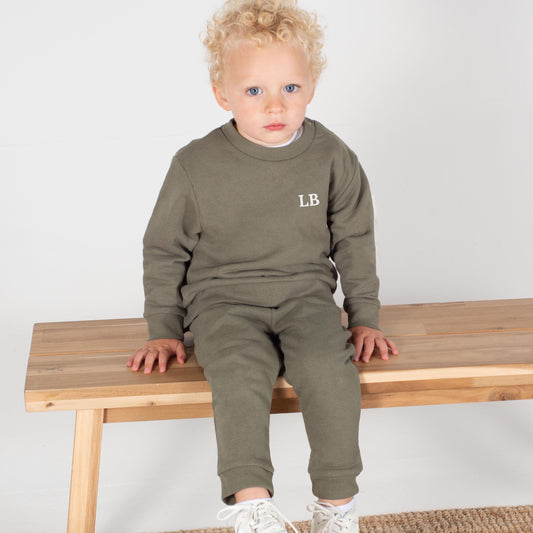 A toddler with blonde curly hair wears a personalised khaki green tracksuit featuring a white printed monogram "LB" on the sweatshirt. The set includes a matching sweatshirt and joggers, both made from soft, eco-friendly cotton-blend fabric. The child is seated on a wooden bench against a neutral backdrop.