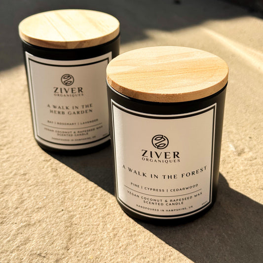 Two scented candles in matte black jars with natural wooden lids, labelled "A Walk in the Forest" and "A Walk in the Herb Garden", positioned in warm natural sunlight on a stone surface.