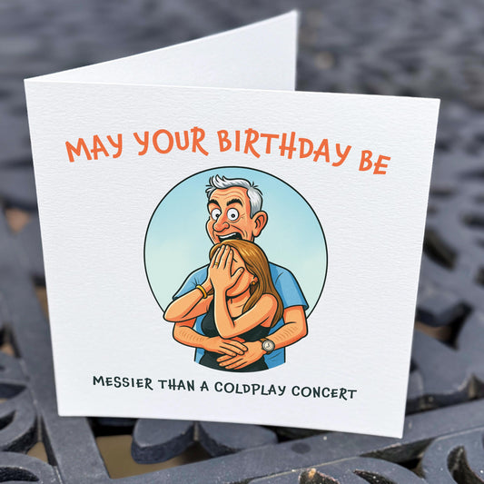 Birthday card featuring a cartoon illustration of a man hugging a woman covering her face, with the text “May your birthday be messier than a Coldplay concert.” Printed on white textured card and shown held above a dark patterned outdoor table.