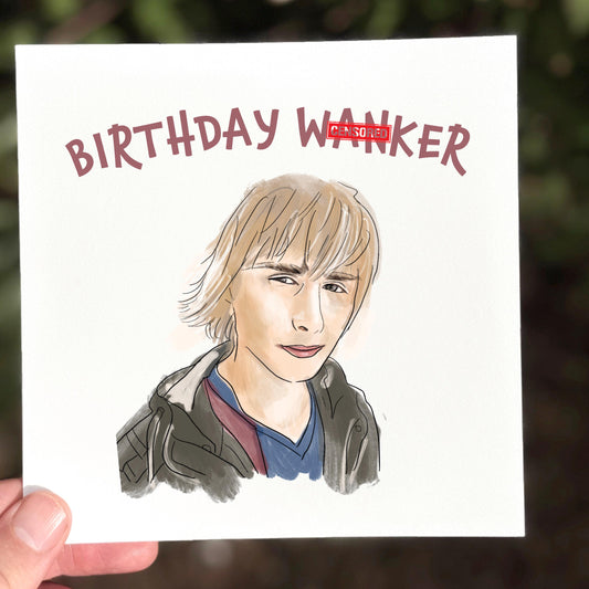 A hand holding a square card featuring a watercolour-style cartoon of a blond young man with the caption “BIRTHDAY WANKER” in bold red lettering above. Printed on off-white textured card and photographed outdoors with a blurred green background.