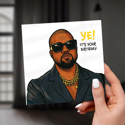 Hand holding a card with a stylized illustration of a person wearing sunglasses and a gold chain, with text 'YE! IT'S YOUR BIRTHDAY'.