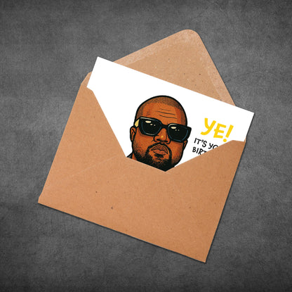 Card with a face design and 'YE! IT'S YOUR BIRTHDAY' text in an open brown envelope on a gray background