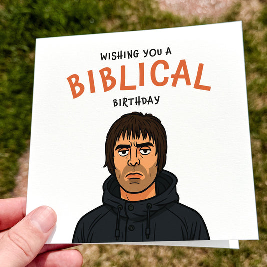 A square white card featuring a cartoon-style drawing of a man with shaggy hair in a black parka, staring deadpan. Above him is the phrase “WISHING YOU A BIBLICAL BIRTHDAY” with “BIBLICAL” in bold orange letters. The design references Britpop music culture. Photographed outdoors on grass in natural light.