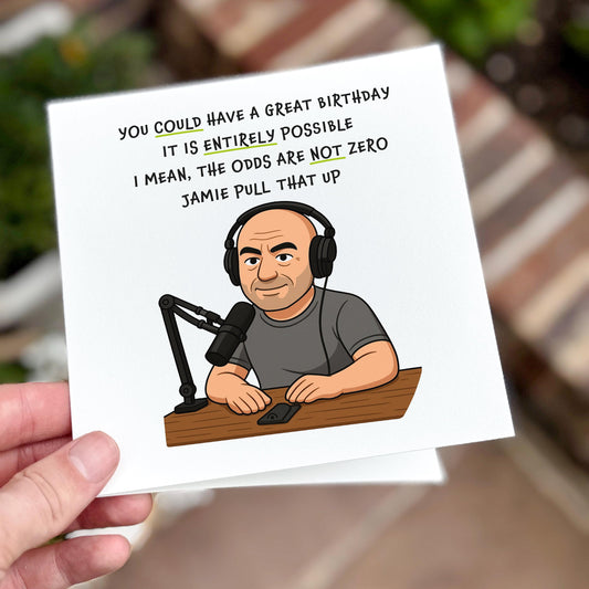 A square birthday card featuring a cartoon-style illustration of a bald man with headphones and a podcast mic setup, mimicking Joe Rogan. Above him, the text reads: “You could have a great birthday. It is entirely possible. I mean, the odds are not zero. Jamie, pull that up.” The tone is dry and meme-driven. Photographed outdoors with a blurred background.