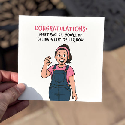 A square congratulations card featuring a cartoon-style woman with a pink top and denim overalls waving cheerfully. The text reads: “CONGRATULATIONS! Meet Rachel, you’ll be seeing a lot of her now.” The tone is funny and highly relatable for new or soon-to-be parents. Photographed outdoors with soft lighting and a blurred background.