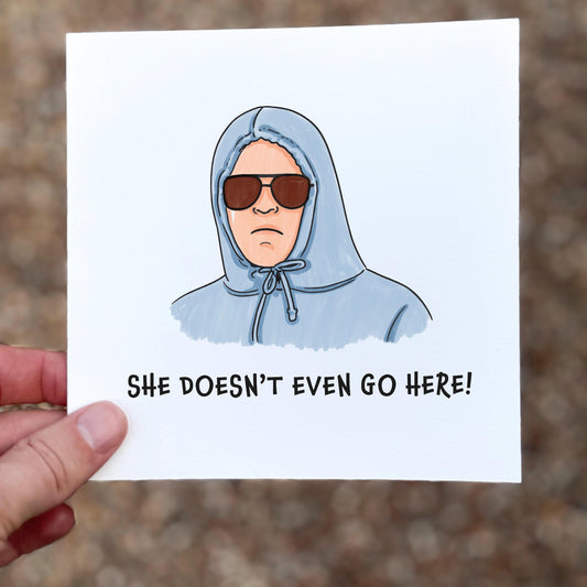 Square illustrated card featuring a cartoon figure in a pale blue hoodie with drawstrings pulled tight, wearing dark sunglasses. Text below reads “SHE DOESN’T EVEN GO HERE!” in bold black font. White background with a minimal, meme-inspired design and sarcastic tone.