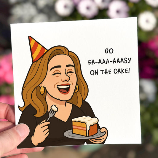 Square illustrated card featuring a cartoon woman resembling Adele, wearing a party hat and laughing while holding a slice of cake and a fork. Text reads “GO EA-AAA-AAASY ON THE CAKE!” in bold black font. The background is white and the tone is light-hearted and musical.