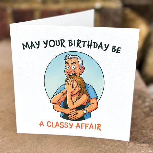Funny birthday card with cartoon illustration of a man looking shocked while hugging a woman covering her face. Text reads “May your birthday be a classy affair” in black and orange. Printed on white textured card and photographed outdoors on stone.