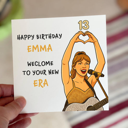 A bold square birthday card featuring a cartoon-style illustration of a female pop star in a glittery gold outfit holding a microphone and forming a heart shape with her hands. The card reads “HAPPY BIRTHDAY EMMA, WELCOME TO YOUR NEW ERA” with the number 13 in metallic gold text in the top right corner, set against a clean white
