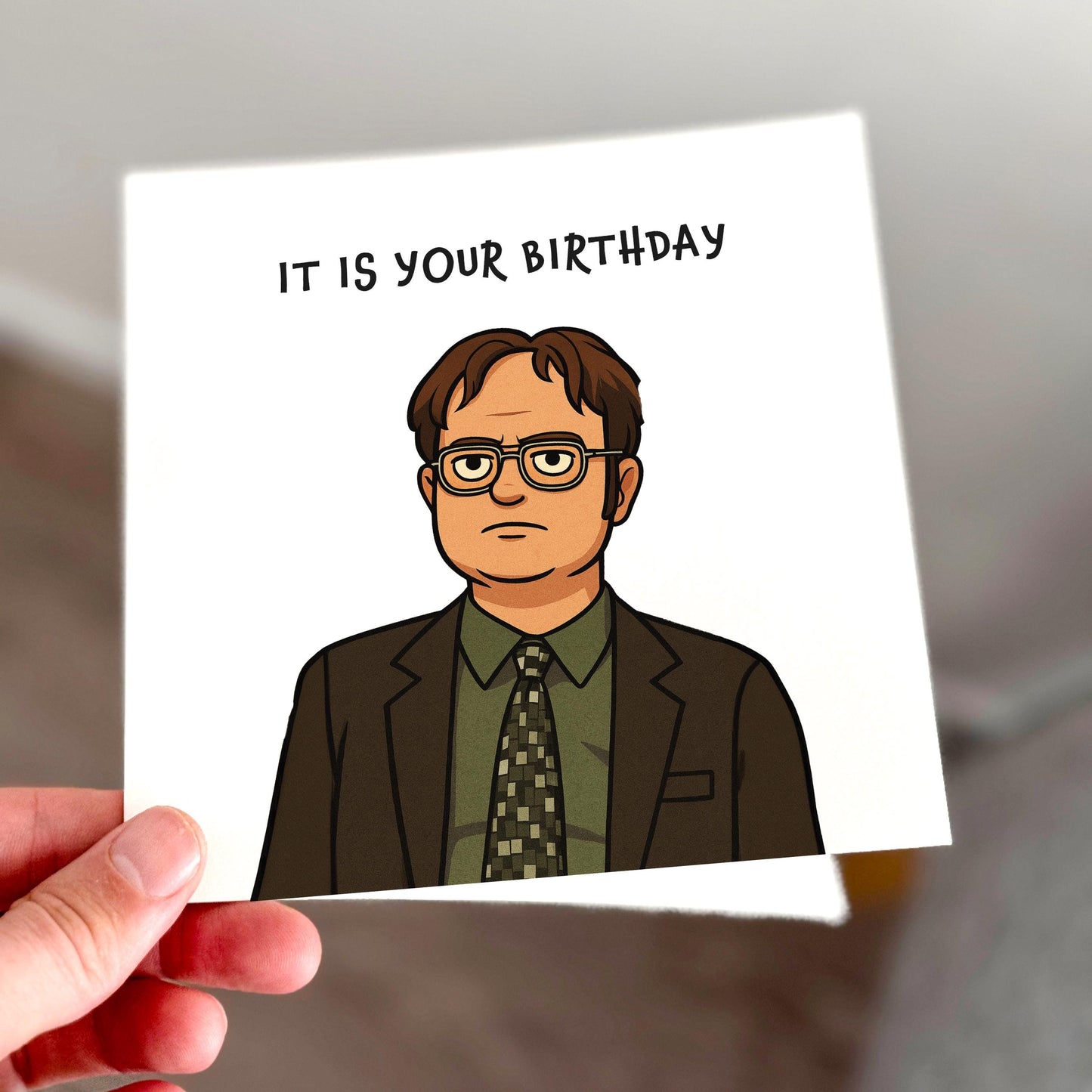 A square birthday card featuring a cartoon-style Dwight Schrute in a green shirt and tie, with the phrase “It is your birthday” printed in plain black text above. The design mimics his famously joyless party planning style from The Office. Card is printed on off-white textured stock and held in hand against a soft background.