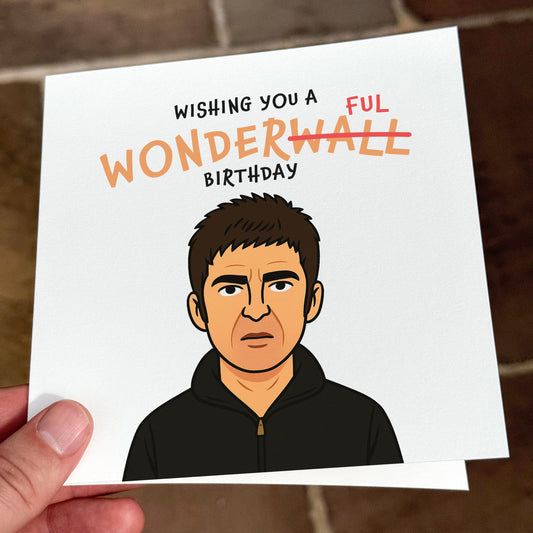 A square birthday card featuring a cartoon drawing of a grumpy-looking man with short dark hair and a black zip-up hoodie. Above him, the text says “WISHING YOU A WONDERFUL BIRTHDAY,” with “WALL” in “WONDERWALL” crossed out and replaced with “FUL” in red. The design references a famous British musician and is shot on a tiled floor background in natural light.