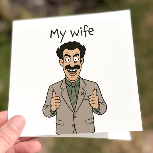A square white greeting card featuring a cartoon illustration of a man in a beige suit with bushy hair and moustache, grinning and giving two thumbs up. Above him, the text reads: “My wife” in a simple black font. The card references a well-known comedy catchphrase. Photographed outdoors with a soft focus green background.