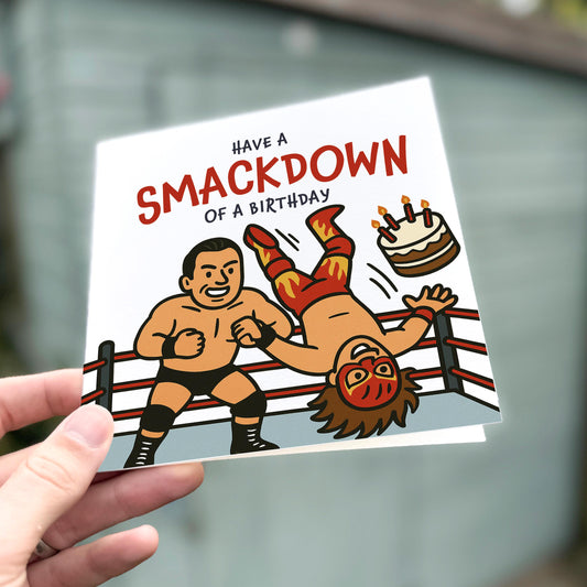 Birthday card with cartoon wrestlers and cake, held by a hand.