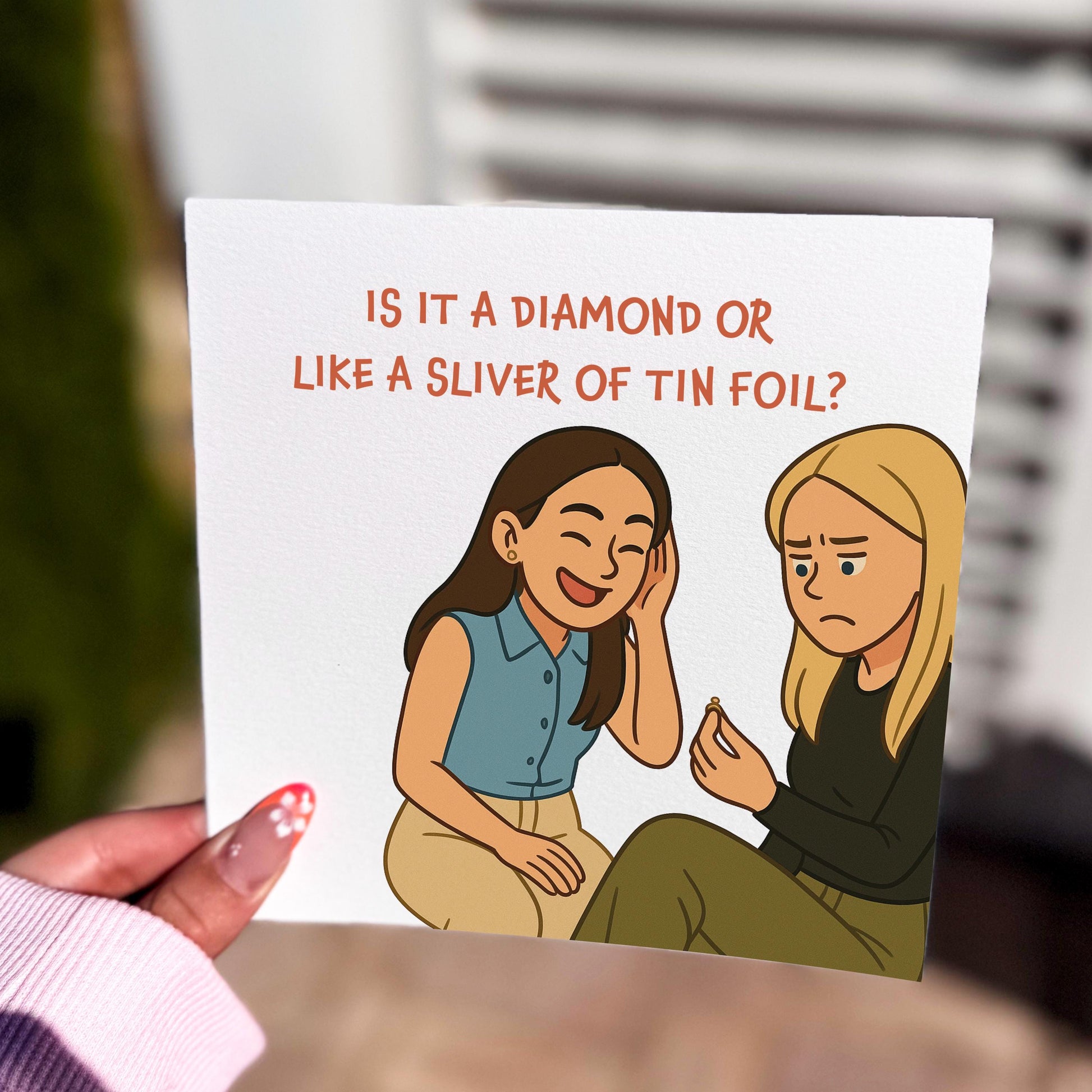 Square greeting card held indoors with a cartoon illustration of two women sitting together. One woman with dark hair is laughing, while the blonde woman looks unimpressed, holding up a ring. Above them, bold red text reads: “IS IT A DIAMOND OR LIKE A SLIVER OF TIN FOIL?” The design is humorous, cheeky, and based on a popular TV show quote.