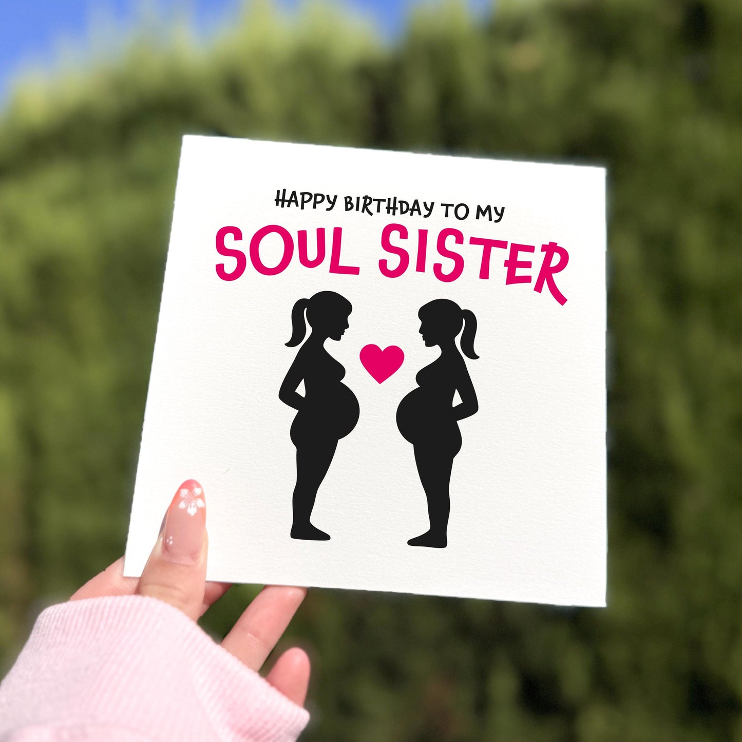 Square greeting card held outdoors in natural light showing two pregnant women in black silhouette facing each other with a pink heart between them. Above them is the text “HAPPY BIRTHDAY TO MY” in black and “SOUL SISTER” in bold pink. A hand with pale pink nails and a pink sleeve holds the card against a blurred green background.