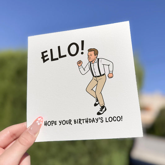 Square greeting card held outdoors in bright natural light. The card shows a cartoon man in beige trousers, suspenders and white shirt doing a quirky dance pose. Above him is the text “ELLO!” and below, “HOPE YOUR BIRTHDAY’S LOCO!” Background is a blurred blue sky and greenery.