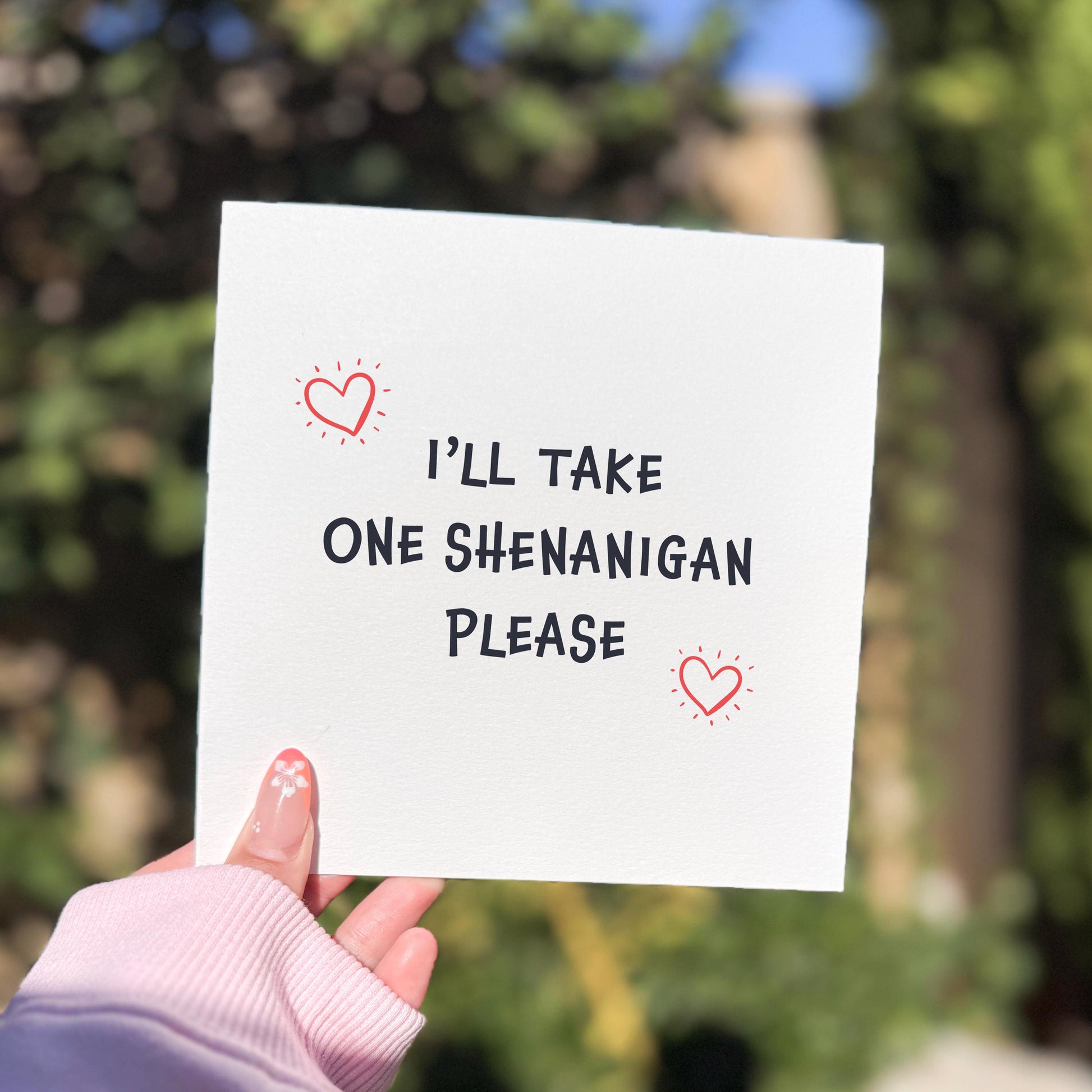 Square greeting card with playful black text reading “I’LL TAKE ONE SHENANIGAN PLEASE” surrounded by two small red hand-drawn hearts. The card is held by a hand wearing a pale pink sleeve, photographed outdoors with a softly blurred green background and bright natural light.