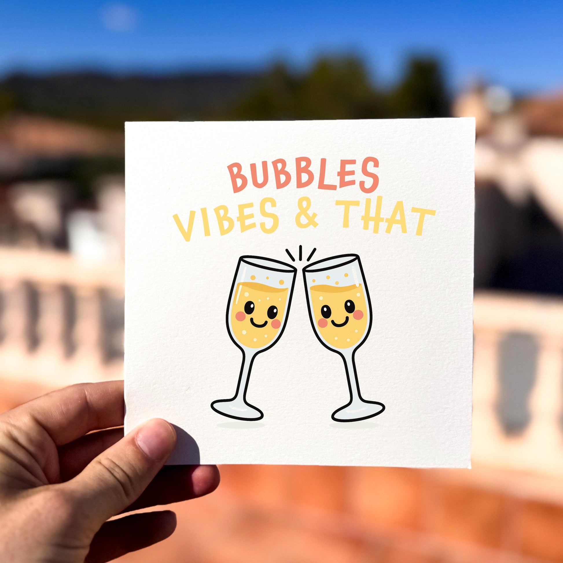 Square greeting card showing two cartoon champagne glasses with smiling faces clinking together, with soft yellow fizz inside. Above them is the text “BUBBLES VIBES & THAT” in pink and yellow lettering. The card is held outdoors in bright sunlight against a blurred terracotta and blue-sky background.