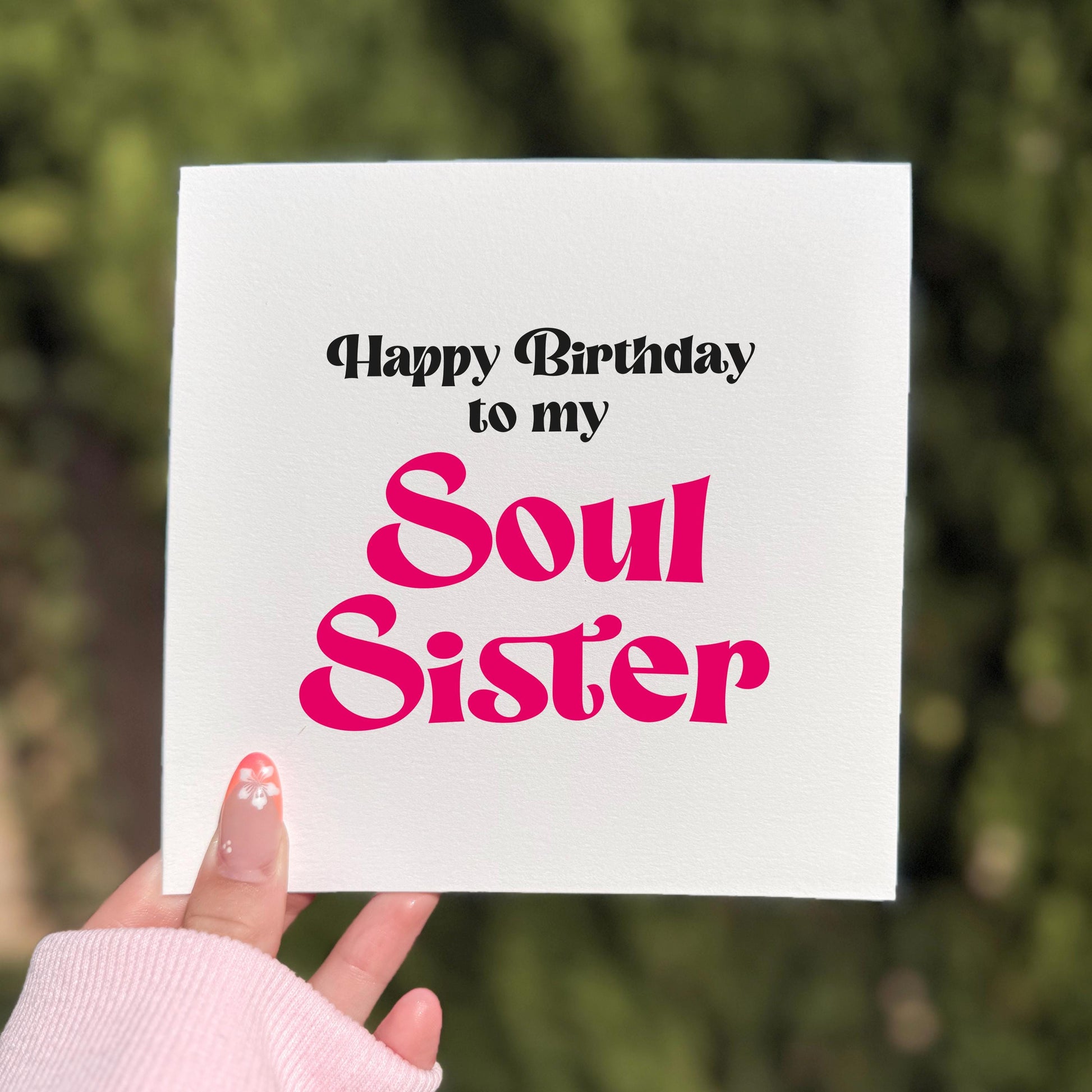 Square greeting card held outdoors in natural light, showing bold black text reading “Happy Birthday to my” above large bright pink retro-style lettering that says “Soul Sister.” A hand with soft pink sleeves and a floral nail design holds the card against a blurred green background.