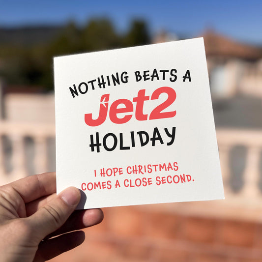 Square Christmas card held outdoors in bright sunlight. The card reads “NOTHING BEATS A Jet2 HOLIDAY” in bold coral and black lettering, with smaller red text underneath saying “I HOPE CHRISTMAS COMES A CLOSE SECOND.” Background shows terracotta rooftops and a blue sky, softly blurred.