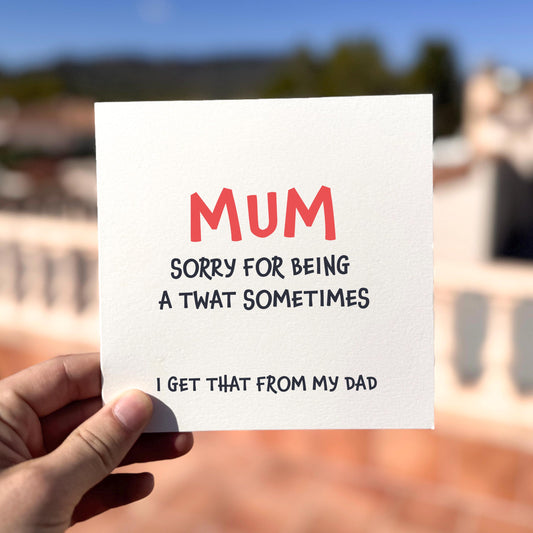 Square greeting card held outdoors in bright sunlight. The card reads “MUM” in large coral text, followed by “SORRY FOR BEING A TWAT SOMETIMES” in black, and smaller text beneath saying “I GET THAT FROM MY DAD.” Background shows a soft blur of terracotta rooftops and blue sky.
