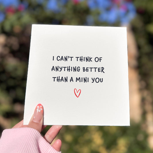 Square greeting card held outdoors in soft natural light. The card reads “I CAN’T THINK OF ANYTHING BETTER THAN A MINI YOU” in simple black lettering, with a small red heart beneath the text. A hand with pink sleeves and floral nail art holds the card against a blurred green and blue background.