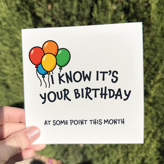Square greeting card held outdoors in bright natural light. The card shows a cluster of colourful balloons (red, orange, yellow, and green) above the text “I KNOW IT’S YOUR BIRTHDAY” and smaller text below reading “AT SOME POINT THIS MONTH.” Background is a softly blurred green hedge.