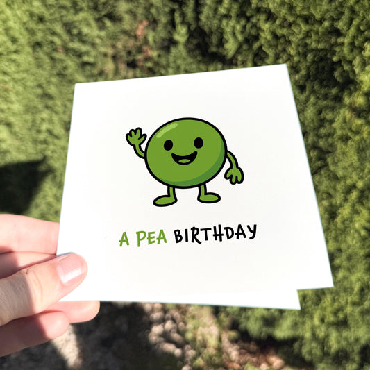 Square greeting card held outdoors in bright natural light. The card features a smiling cartoon green pea character waving, with the text “A PEA BIRTHDAY” underneath in mixed green and black lettering. Background is a softly blurred hedge.
