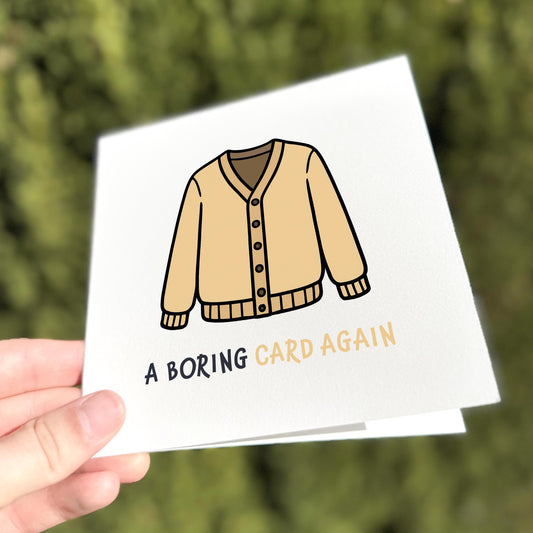 Square greeting card held outdoors in bright natural light. The design shows a beige button-up cardigan illustration, with the text “A BORING CARD AGAIN” beneath it in mixed black and beige lettering. Background is a softly blurred hedge.