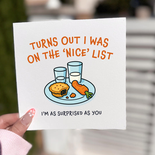 Square Christmas card held outdoors in soft natural light. The card shows a plate with a mince pie, crumbs, a carrot and two glasses of milk. Above it reads “TURNS OUT I WAS ON THE ‘NICE’ LIST,” with “I’M AS SURPRISED AS YOU” below. Background is softly blurred greenery and white fencing.