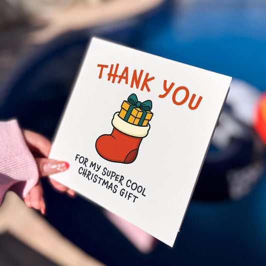 Square Christmas card held outdoors in bright natural light. The design shows a red stocking filled with wrapped presents, with the text “THANK YOU” above and “FOR MY SUPER COOL CHRISTMAS GIFT” below in playful lettering. Background includes a blurred pool and hand with pink sleeves.